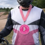 BMX rider wearing a pink breast cancer awareness race medallion with a first place design, featuring Race For Breast Cancer and Snake River BMX text on a pink ribbon, custom medallion by BML Trophies and Awards.